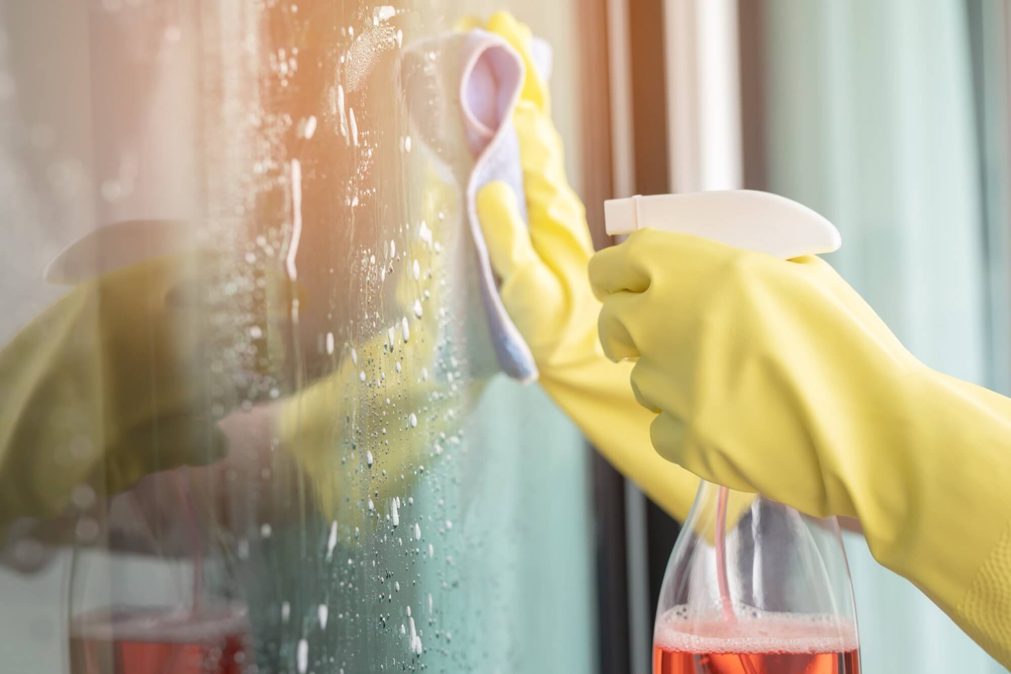 A person wearing yellow gloves sprays detergent and wipes a window with a cloth.